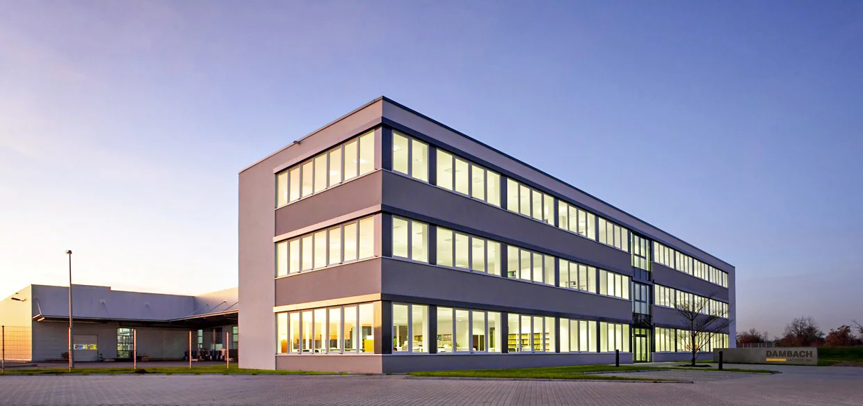 Modernes mehrstöckiges DAMBACH Bürogebäude mit großflächigen Fenstern und beleuchteten Innenräumen bei Dämmerung.
